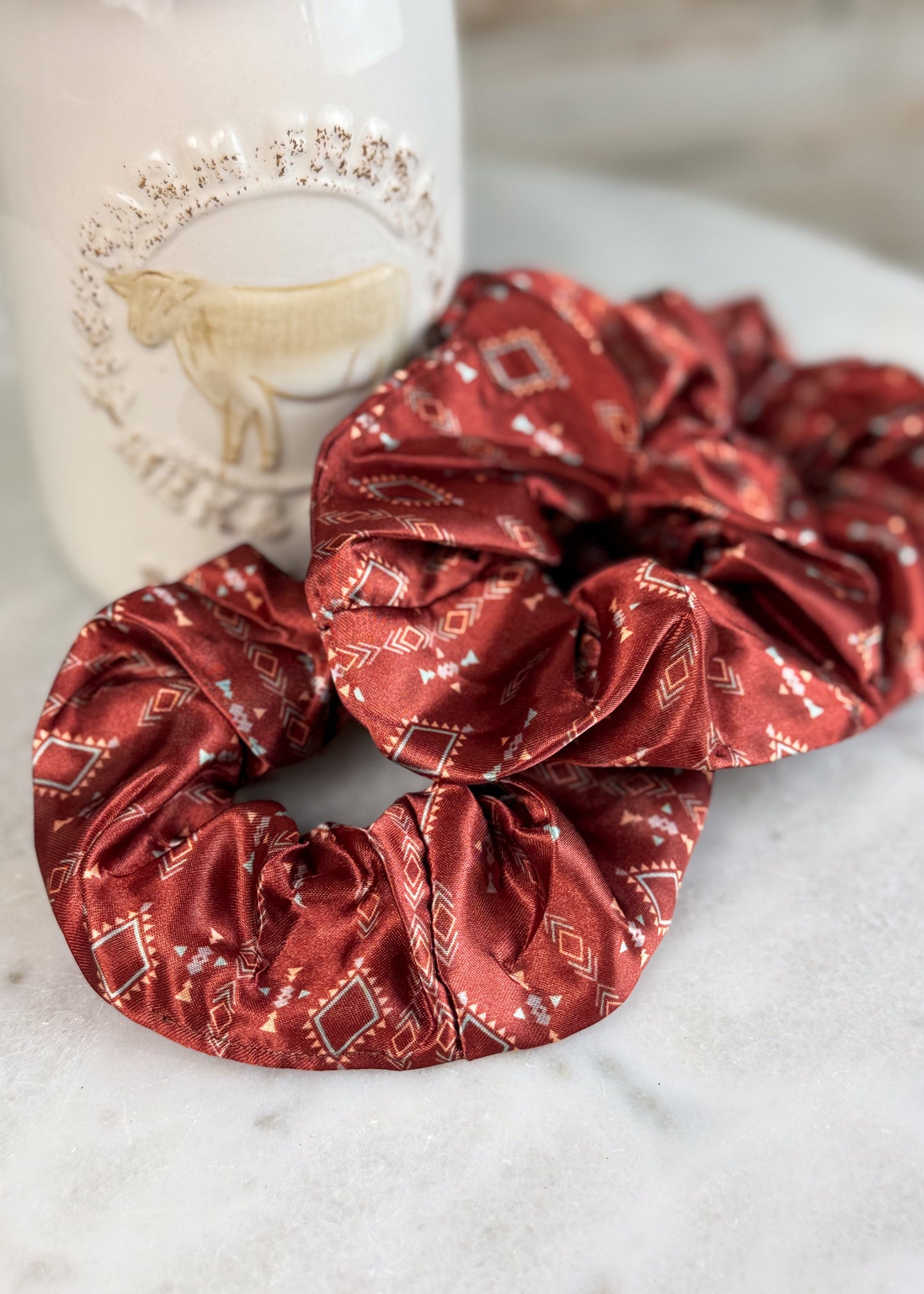 Two red satin hair scrunchies with a white geometric pattern, displayed on a light marble surface with a white ceramic mug in the background.