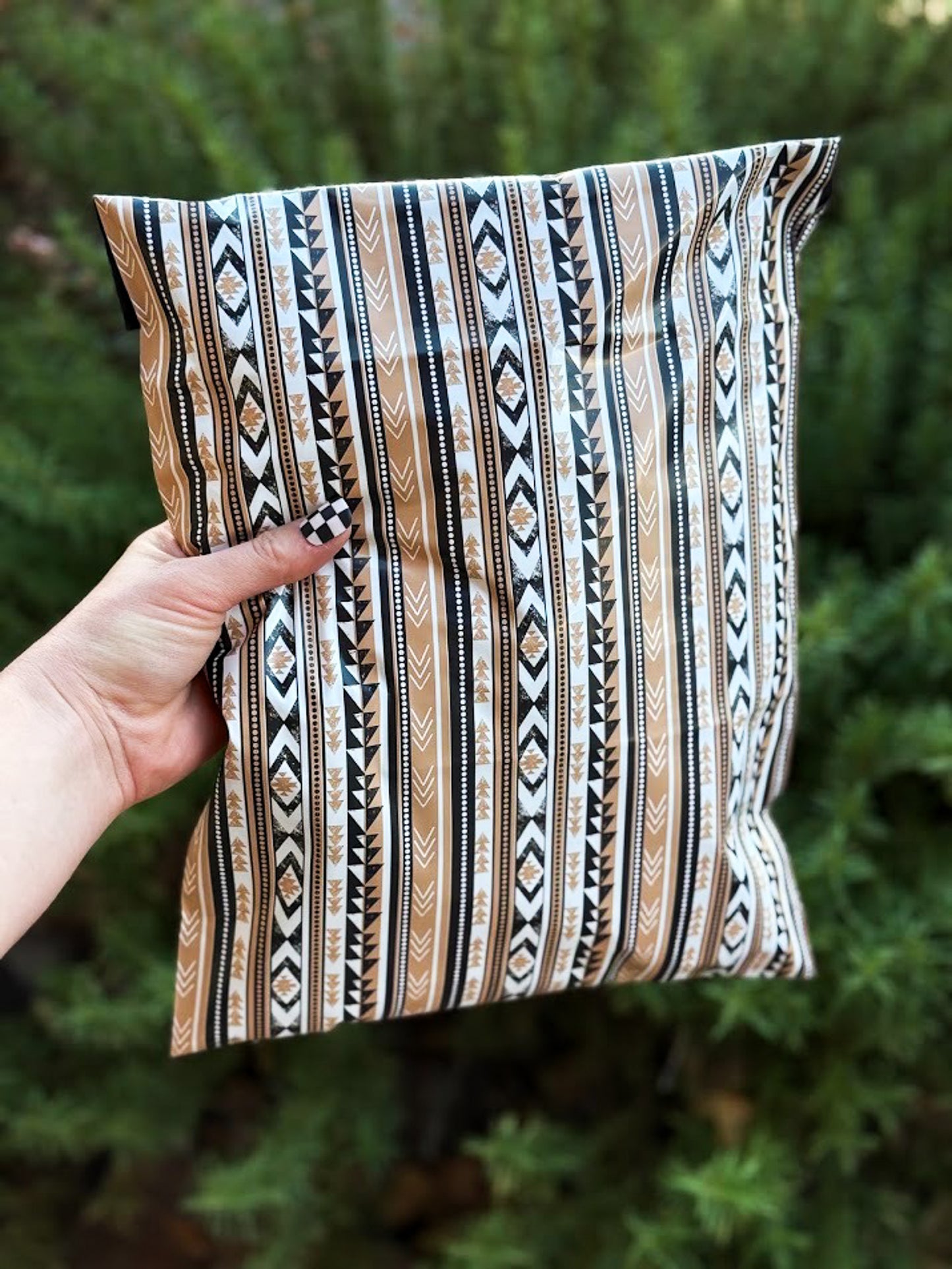 A hand holding as shipping poly mailer with a vertical Southwestern-style geometric pattern in black, white, and tan, set against a blurred green outdoor 