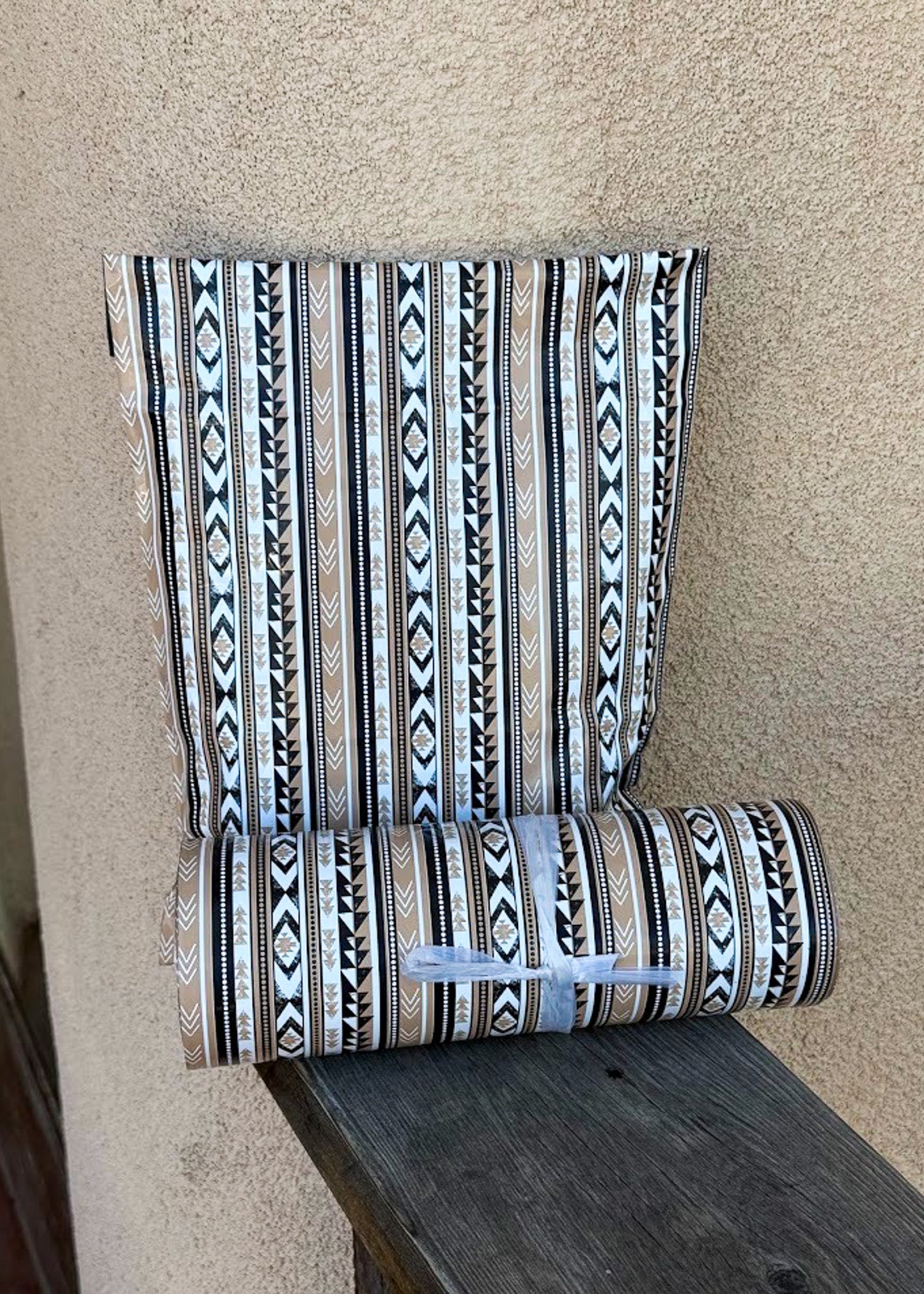 A roll of patterned wallpaper or fabric with vertical geometric tribal stripes in black, white, beige, and brown, partially unrolled and resting on a dark wooden surface against a textured beige wall.