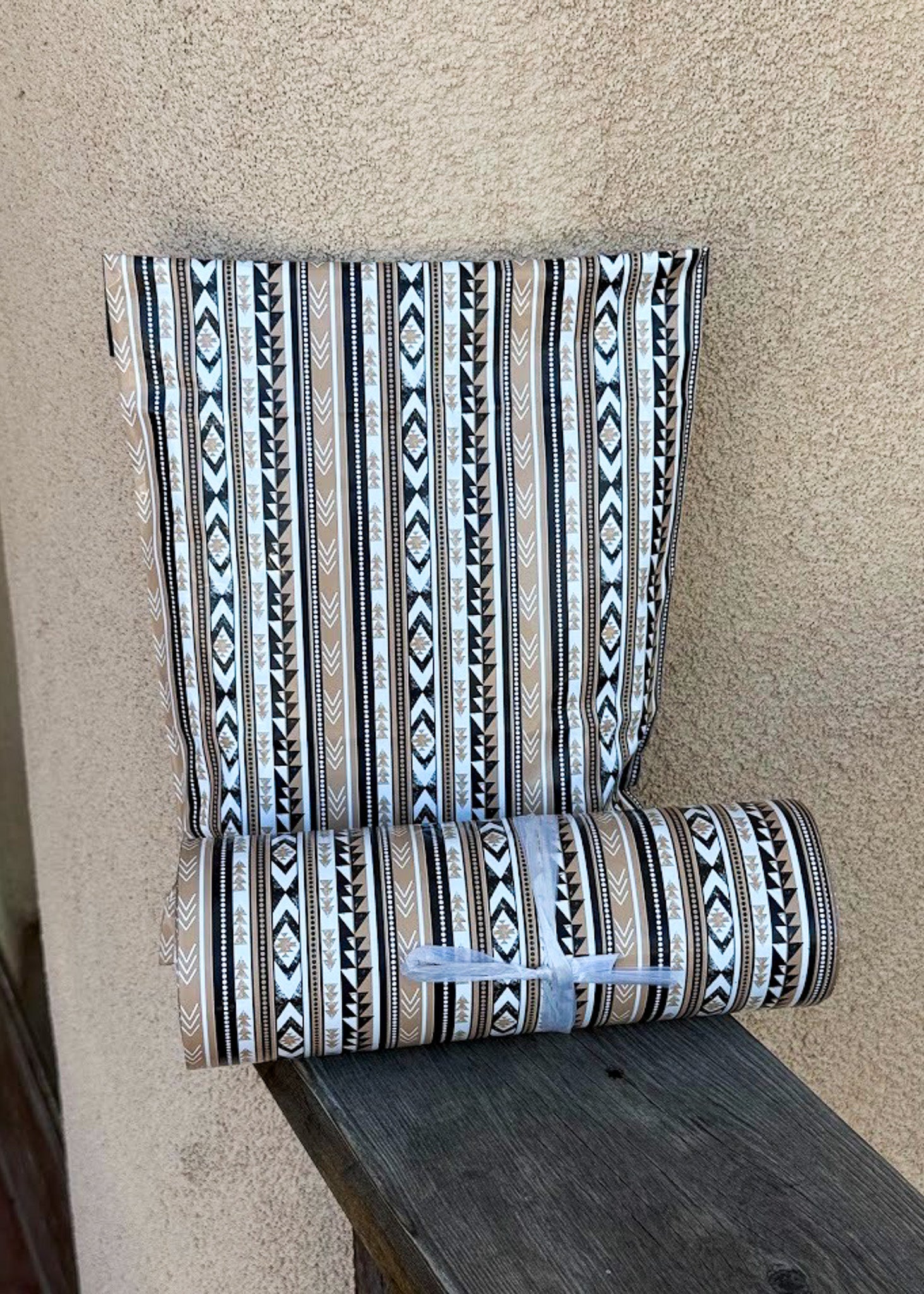 A roll of patterned wallpaper or fabric with vertical geometric tribal stripes in black, white, beige, and brown, partially unrolled and resting on a dark wooden surface against a textured beige wall.