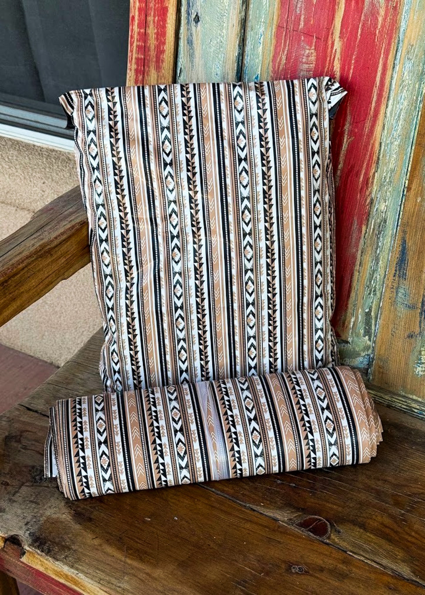 A folded and upright view of a patterned poly mailer bag on a rustic wooden porch. The cushion features a Southwestern-style geometric stripe pattern in beige, brown, black, and muted orange.