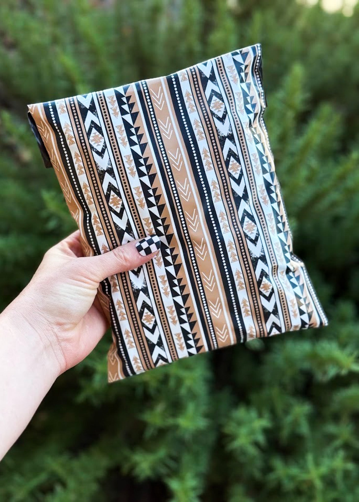 A hand holding a poly mailer with a vertical Southwestern-style geometric pattern in beige, black, and white, set against a blurred background of green foliage.