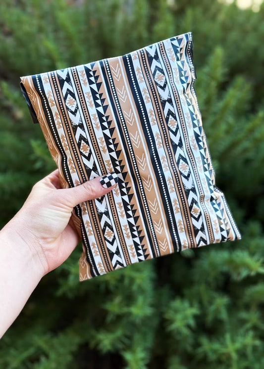 A hand holding a poly mailer with a vertical Southwestern-style geometric pattern in beige, black, and white, set against a blurred background of green foliage.