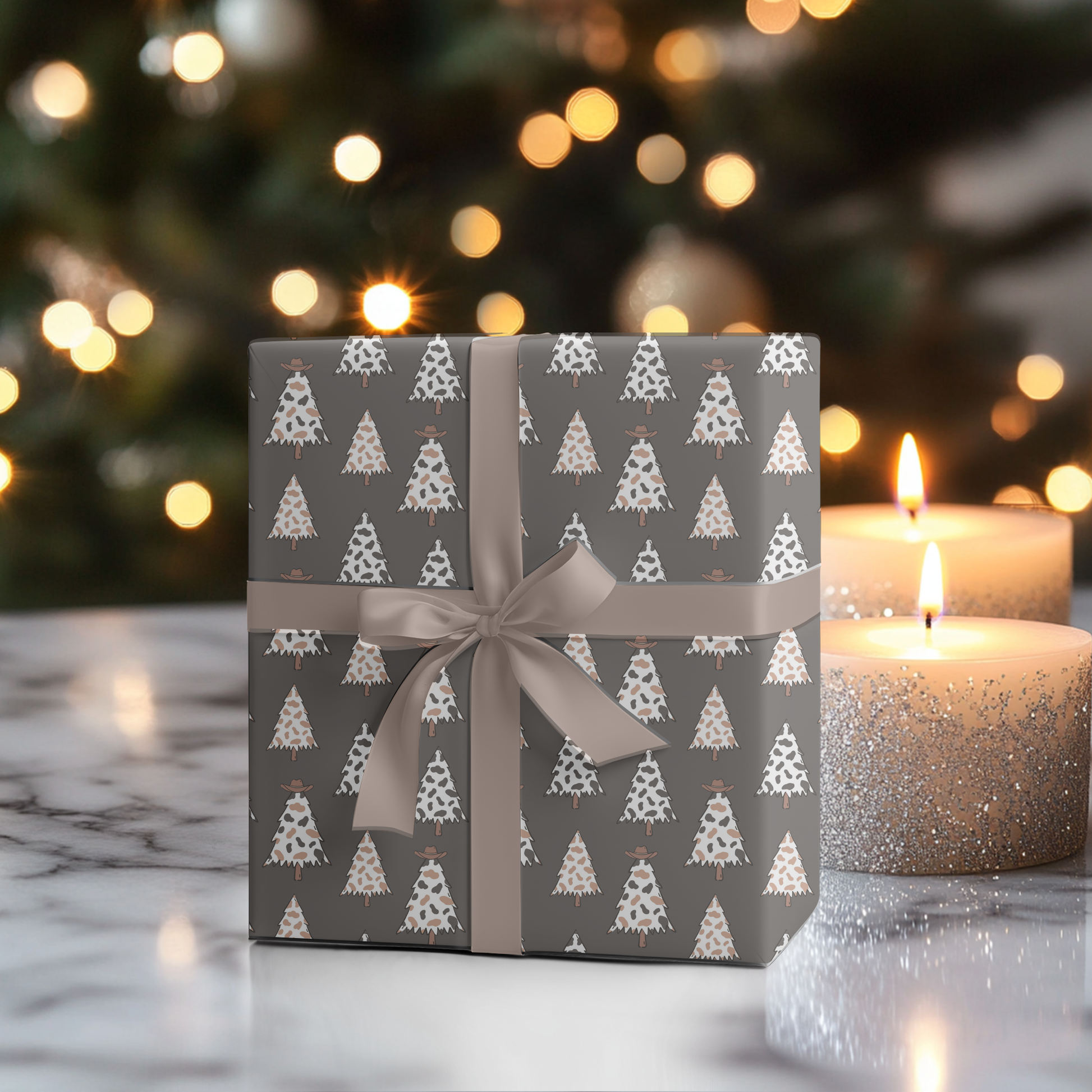 A square gift box wrapped in gray paper with a white Christmas tree pattern and tied with a taupe ribbon, set on a marble surface with lit candles and a blurred Christmas tree in the background.