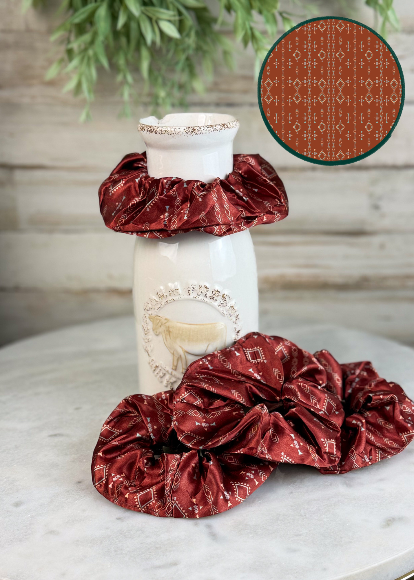 Two burgundy satin hair scrunchies with a white geometric pattern, displayed on a white ceramic vase and on a marble surface. A circular inset in the top right shows a close-up of the fabric pattern.